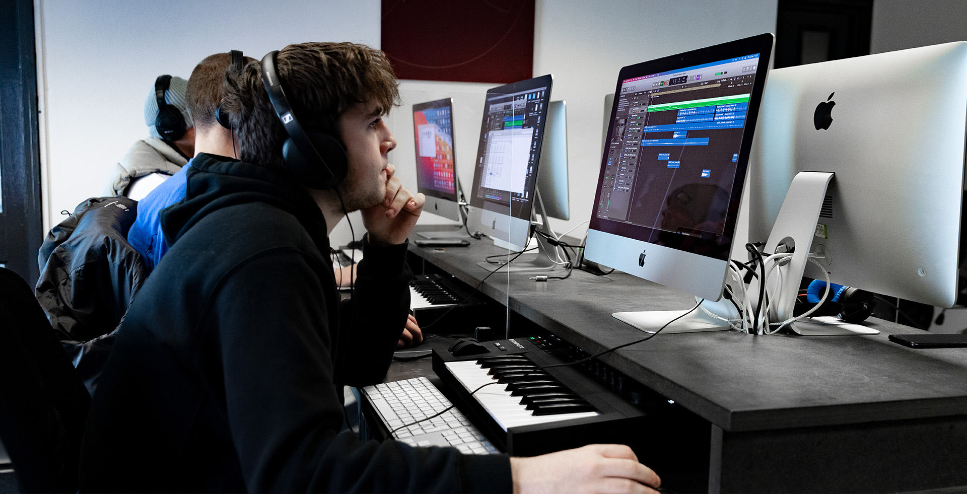 college student with headphones using an apple mac to produce music. There is a keyboard on his desk