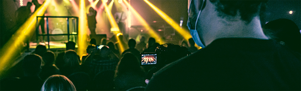 A male filming a band performing and an audience watching from the back of a venue